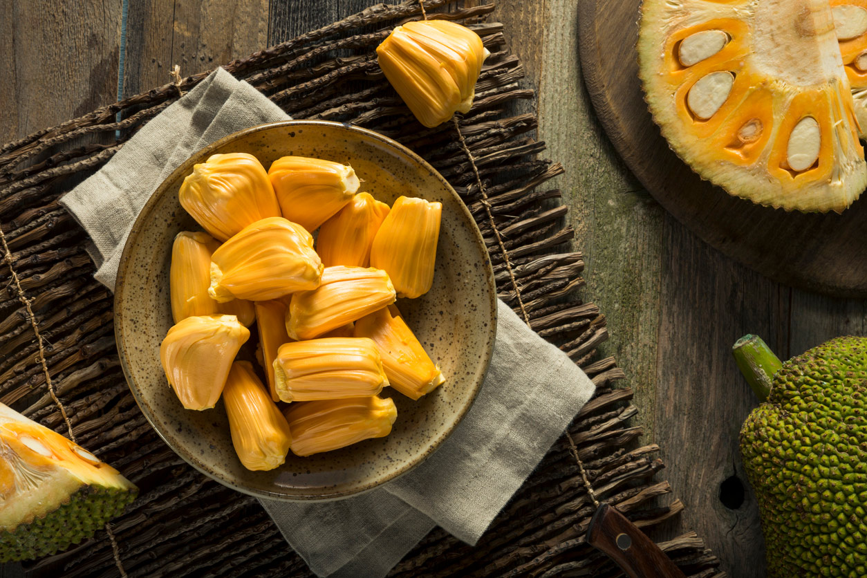 Jackfruit pods in a bowl on a wooden table next to a halved jackfruit with pod segments visible